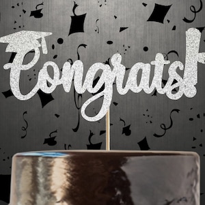 Pode incluir: Topper de bolo de glitter prateado com a palavra "Parabéns!" e um chapéu de formatura no topo. O topper está em um bolo preto.