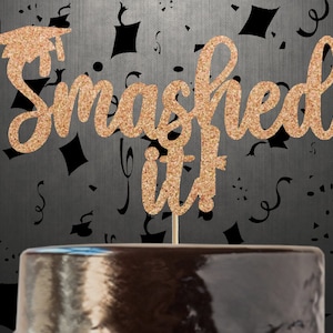 Pode incluir: Topper de bolo de glitter dourado com o texto "Smashed it!" e um chapéu de formatura. O topper está em um fundo preto com confete preto.