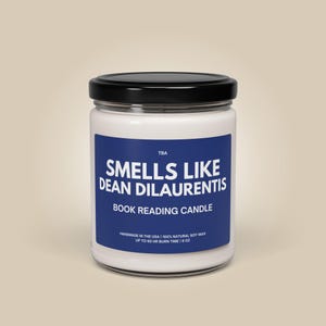 May include: A white glass jar candle with a black lid. The candle has a blue label with the text "SMELLS LIKE DEAN DILAURENTIS BOOK READING CANDLE". The label also includes the text "HANDMADE IN THE USA | 100% NATURAL SOY WAX UP TO 60 HR BURN TIME | 9 OZ".
