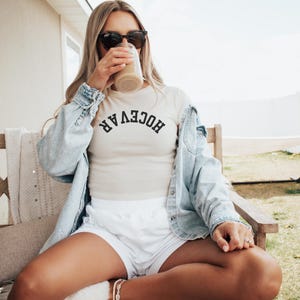 Puede incluir: Un top corto color crema con la palabra "HOCEVAR" en letras negras arqueadas. La persona lleva una chaqueta vaquera azul claro, pantalones cortos blancos y gafas de sol negras. La persona está bebiendo de un vaso transparente.