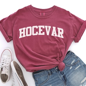 Puede incluir: Una camiseta de color granate con la palabra "HOCEVAR" impresa en blanco en la parte delantera. La camiseta está colocada sobre una superficie blanca con un par de zapatillas blancas y unos pantalones cortos de mezclilla azul.