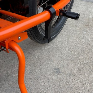 K&ouml;nnte beinhalten: Orangener Fahrrad-Lastanh&auml;nger mit schwarzem Rad und schwarzem St&auml;nder. Der Anh&auml;nger hat einen schwarzen Metallrahmen und ein schwarzes Fu&szlig;pedal.