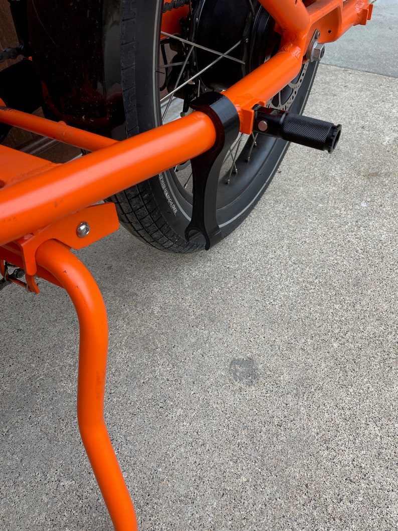 K&ouml;nnte beinhalten: Orangener Fahrrad-Lastanh&auml;nger mit schwarzem Rad und schwarzem St&auml;nder. Der Anh&auml;nger hat einen schwarzen Metallrahmen und ein schwarzes Fu&szlig;pedal.