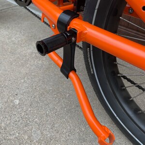K&ouml;nnte beinhalten: Nahaufnahme eines orangefarbenen Fahrradrahmens mit einem schwarzen St&auml;nder. Der St&auml;nder ist mit einer schwarzen Metallhalterung am Rahmen befestigt. Der St&auml;nder ist ausgeklappt und das Fahrrad steht auf einer Betonfl&auml;che.