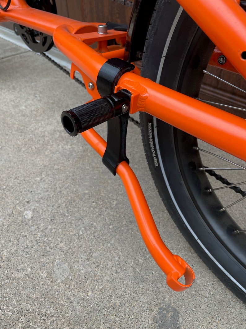 K&ouml;nnte beinhalten: Nahaufnahme eines orangefarbenen Fahrradrahmens mit einem schwarzen St&auml;nder. Der St&auml;nder ist mit einer schwarzen Metallhalterung am Rahmen befestigt. Der St&auml;nder ist ausgeklappt und das Fahrrad steht auf einer Betonfl&auml;che.