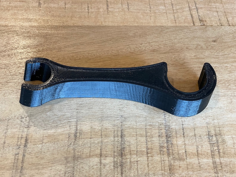 K&ouml;nnte beinhalten: Ein schwarzes Plastikwerkzeug mit zwei Schlaufen an beiden Enden. Das Werkzeug ist zum &Ouml;ffnen und Schlie&szlig;en von Flaschenverschl&uuml;ssen gedacht.