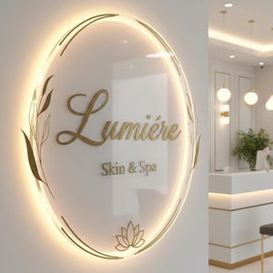 Może przedstawiać: Podświetlany szyld dla "Lumière Skin & Spa" ze złotymi literami i złotą owalną ramą. Znak zawiera kwiat lotosu i akcenty botaniczne. Tło przedstawia recepcję spa z białym i złotym wystrojem.