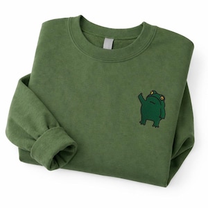May include: Olive green sweatshirt with a crew neck and long sleeves. A dark green embroidered frog graphic is on the front. The sweatshirt is folded, showing the cuff of the sleeve.