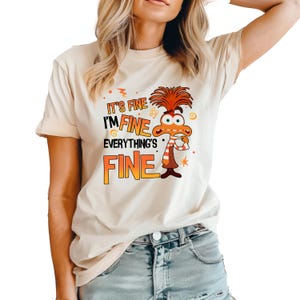 May include: A beige t-shirt with a cartoon character and the text "It's fine I'm fine everything's fine".