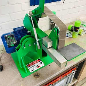 Puede incluir: Una lijadora de banda verde con un motor eléctrico azul y una palanca de seguridad. La lijadora está sobre un banco de trabajo de madera con una caja de almacenamiento de plástico gris. La lijadora tiene una señal de advertencia que dice "Danger Engage Safety Squint".