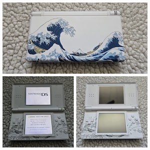 May include: White Nintendo DS handheld game console with a custom design featuring the iconic "Great Wave off Kanagawa" artwork. The console is open and closed, showing the screens and the wave design.