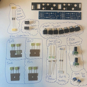 May include: A collection of electronic components for a modular synthesiser kit, including a black faceplate, a blue circuit board, mono jacks, DIP switches, resistors, film capacitors, green LEDs, LDRs, diodes, potentiometers, and trim pots.