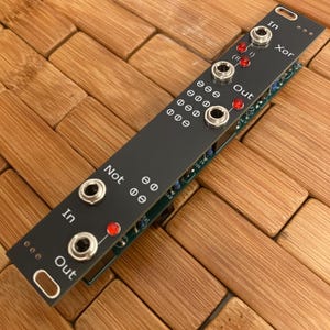 May include: A black modular synthesizer panel featuring silver audio jacks and red LED indicators. The panel is labeled with white text including In, Out, Xor, and Not, along with various logic symbols and circuit board components visible underneath.