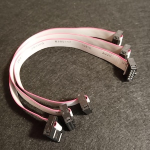 Pode incluir: Três cabos de fita plana com conectores. Os cabos são brancos com uma faixa rosa e conectores pretos. O texto no cabo diz "28AWG", "E390147" e "105C". Os conectores são pretos e brancos.