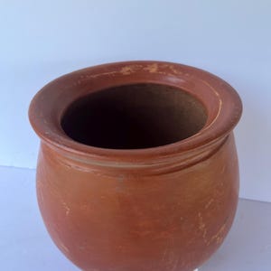 May include: A terracotta-colored, round ceramic pot with a wide opening and a slightly textured surface. The pot has a rounded body and a thick rim. The interior is dark brown. The pot is likely used for planting or storage.