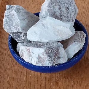 K&ouml;nnte beinhalten: Eine Sammlung hellgrauer, unregelm&auml;&szlig;ig geformter Steine in einer kleinen, runden, blauen Schale. Die Steine haben eine strukturierte Oberfl&auml;che und verschiedene Grau- und Wei&szlig;t&ouml;ne. Die Schale steht auf einer Holzoberfl&auml;che.