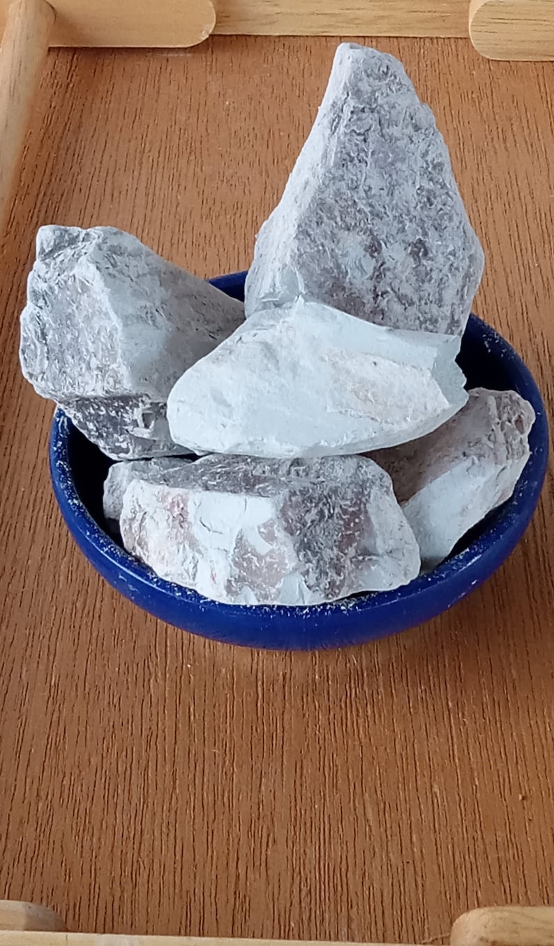 K&ouml;nnte beinhalten: Eine Sammlung hellgrauer, unregelm&auml;&szlig;ig geformter Steine in einer kleinen, runden, blauen Schale. Die Steine haben eine strukturierte Oberfl&auml;che und verschiedene Grau- und Wei&szlig;t&ouml;ne. Die Schale steht auf einer Holzoberfl&auml;che.