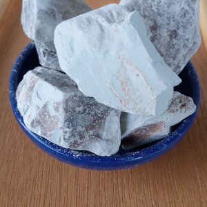 K&ouml;nnte beinhalten: Eine Sammlung hellblauer, unregelm&auml;&szlig;ig geformter Steine ​​f&uuml;llt eine kleine, runde, dunkelblaue Sch&uuml;ssel. Die Steine ​​haben eine raue Textur und einige haben braune und beige Flecken. Die Sch&uuml;ssel steht auf einer Holzoberfl&auml;che.