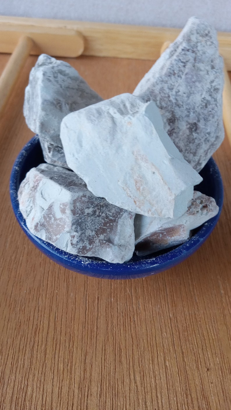 K&ouml;nnte beinhalten: Eine Sammlung hellblauer, unregelm&auml;&szlig;ig geformter Steine ​​f&uuml;llt eine kleine, runde, dunkelblaue Sch&uuml;ssel. Die Steine ​​haben eine raue Textur und einige haben braune und beige Flecken. Die Sch&uuml;ssel steht auf einer Holzoberfl&auml;che.
