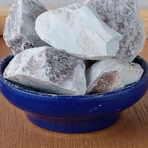 K&ouml;nnte beinhalten: Eine Sammlung hellblauer und grauer Steine, m&ouml;glicherweise Mineralien oder Kristalle, wird in einer kleinen, tiefblauen Keramikschale pr&auml;sentiert. Die Steine haben eine raue, strukturierte Oberfl&auml;che und verschiedene Farbt&ouml;ne. Die Schale steht auf einer Holzoberfl&auml;che.