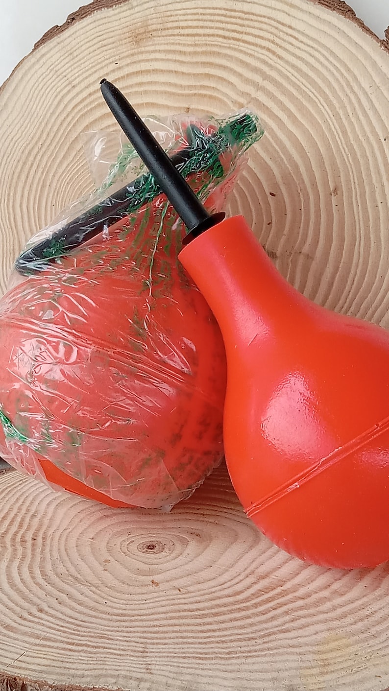 May include: Two red rubber bulb syringes with a black nozzle. The syringes are on a wooden surface.
