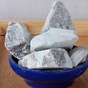 K&ouml;nnte beinhalten: Eine Ansammlung hellgrauer Felsen mit strukturierten Oberfl&auml;chen, pr&auml;sentiert in einer kleinen, runden, kobaltblauen Schale. Die Felsen variieren in Gr&ouml;&szlig;e und Form und erzeugen eine nat&uuml;rliche &Auml;sthetik. Die Schale steht auf einer Holzoberfl&auml;che.