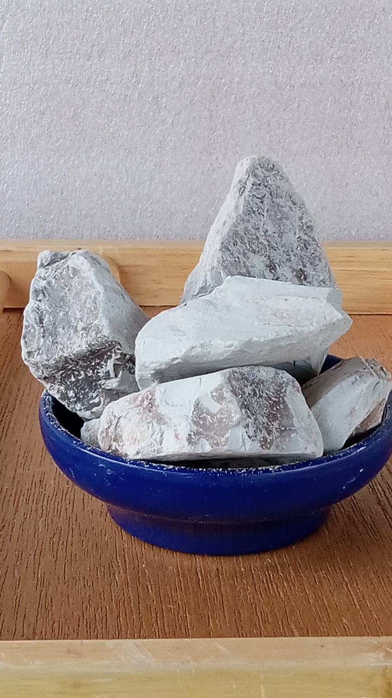 K&ouml;nnte beinhalten: Eine Ansammlung hellgrauer Felsen mit strukturierten Oberfl&auml;chen, pr&auml;sentiert in einer kleinen, runden, kobaltblauen Schale. Die Felsen variieren in Gr&ouml;&szlig;e und Form und erzeugen eine nat&uuml;rliche &Auml;sthetik. Die Schale steht auf einer Holzoberfl&auml;che.