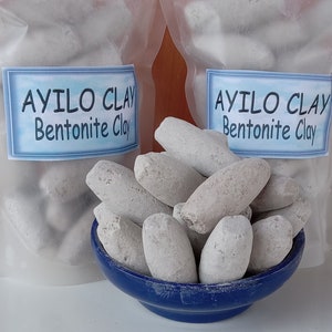 May include: A blue bowl filled with white, oval-shaped bentonite clay stones. The bowl is sitting on a white surface. Two clear plastic bags with the text "AYILO CLAY Bentonite Clay" are in the background.