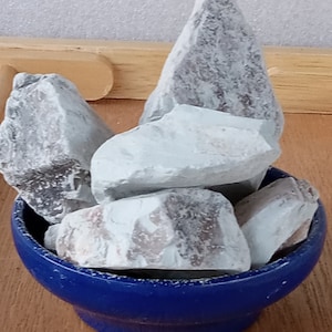 May include: A collection of light blue and gray rocks fills a small, round, dark blue bowl. The rocks have a rough, textured surface. The bowl sits on a wooden surface, with a light gray background.
