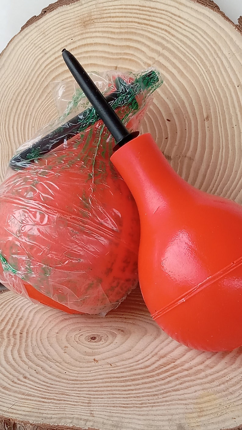 May include: Two red rubber bulb syringes with a black nozzle. The syringes are on a wooden surface.