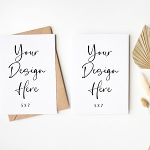 K&ouml;nnte beinhalten: Zwei leere wei&szlig;e Karten mit dem Text "Your Design Here 5x7" auf jeder Karte. Die Karten liegen auf einer wei&szlig;en Oberfl&auml;che mit einem braunen Umschlag und getrockneten Palmbl&auml;ttern und Blumen.