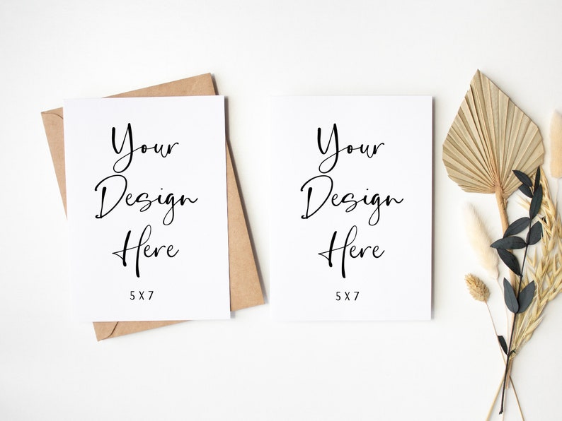 K&ouml;nnte beinhalten: Zwei leere wei&szlig;e Karten mit dem Text "Your Design Here 5x7" auf jeder Karte. Die Karten liegen auf einer wei&szlig;en Oberfl&auml;che mit einem braunen Umschlag und getrockneten Palmbl&auml;ttern und Blumen.