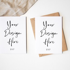 K&ouml;nnte beinhalten: Zwei leere wei&szlig;e Karten mit dem Text "Your Design Here" und "5x7" darauf gedruckt. Die Karten liegen auf einer wei&szlig;en Oberfl&auml;che mit einem braunen Umschlag und beigefarbenem Stoff im Hintergrund.