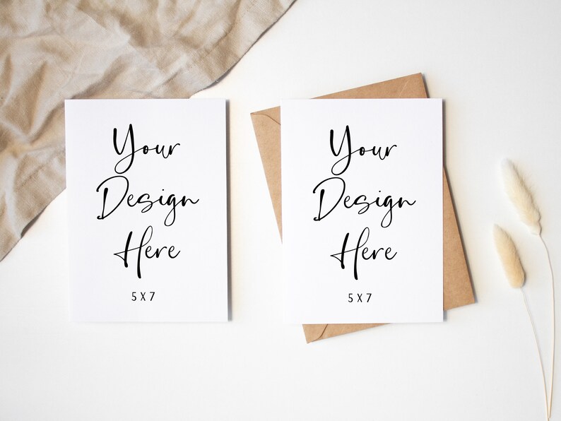 K&ouml;nnte beinhalten: Zwei leere wei&szlig;e Karten mit dem Text "Your Design Here" und "5x7" darauf gedruckt. Die Karten liegen auf einer wei&szlig;en Oberfl&auml;che mit einem braunen Umschlag und beigefarbenem Stoff im Hintergrund.