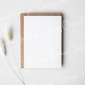 May include: A blank white card with a brown envelope on a white background. Two dried white pampas grass stems are in the lower left corner of the image.