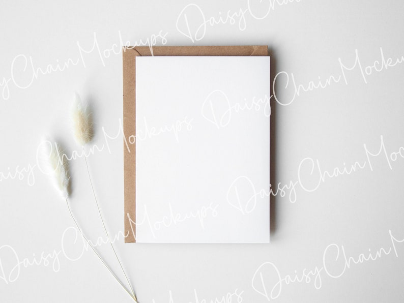 May include: A blank white card with a brown envelope on a white background. Two dried white pampas grass stems are in the lower left corner of the image.