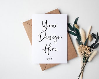 May include: A blank white card with the text "Your Design Here" and "5x7" printed on it. The card is sitting on a brown envelope with dried flowers and greenery next to it.