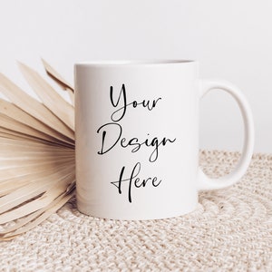 May include: White ceramic mug with a black inscription that reads "Your Design Here" on a beige woven mat with a dried palm leaf.