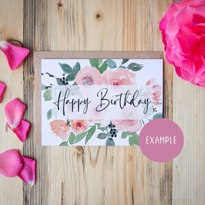 May include: A white card with a watercolor floral design and the text "Happy Birthday" in black script. The card is surrounded by pink rose petals on a wooden background.