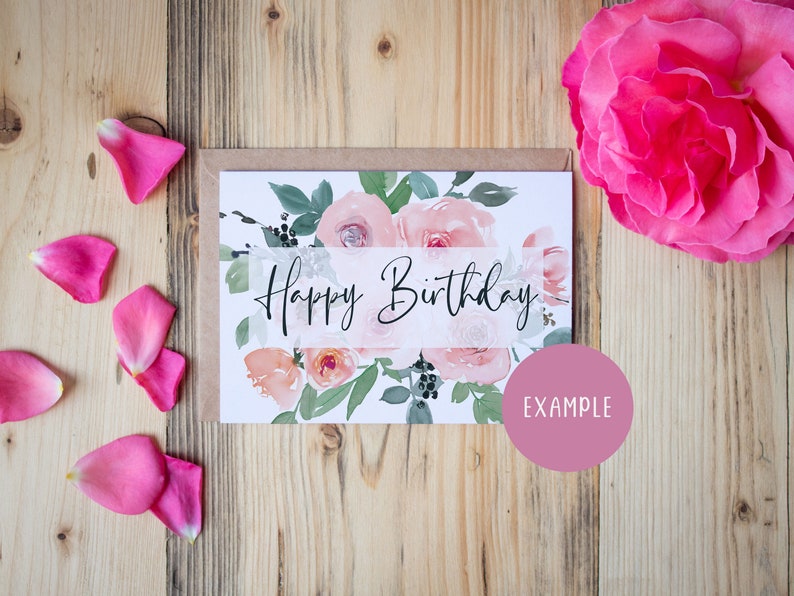 May include: A white card with a watercolor floral design and the text "Happy Birthday" in black script. The card is surrounded by pink rose petals on a wooden background.