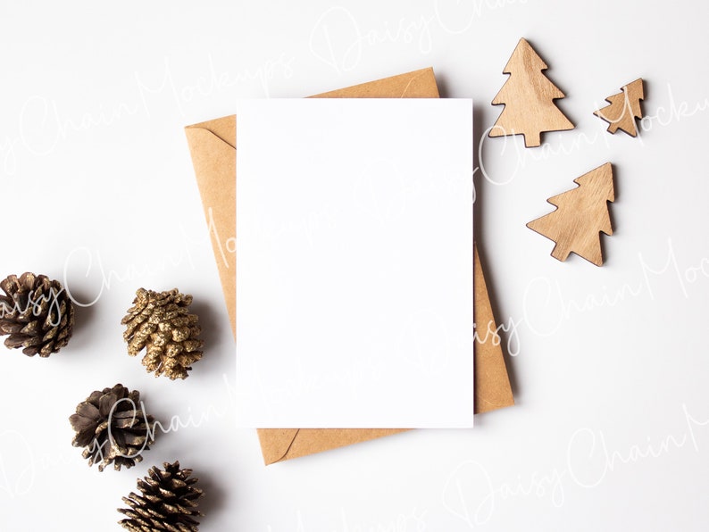 K&ouml;nnte beinhalten: Eine leere wei&szlig;e Karte mit einem braunen Umschlag, Tannenzapfen und h&ouml;lzernen Weihnachtsbaumschmuck auf wei&szlig;em Hintergrund.