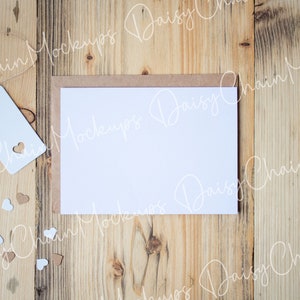 May include: A blank white card with a brown envelope on a wooden background. There are small white and brown hearts scattered around the card.