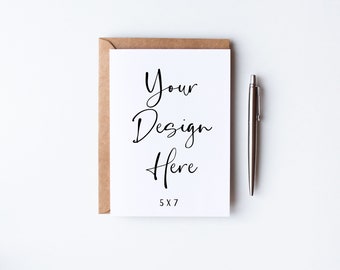 May include: A white card with a brown envelope behind it. The card has the text "Your Design Here" written in black cursive. The card also has the text "5x7" printed in black at the bottom. A silver pen is lying on a white surface to the right of the card.