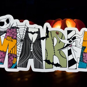 May include: A colorful, patchwork-style name sign that reads "MARIA" with a Jack Skellington character from the Nightmare Before Christmas movie. The sign is decorated with bats, spiderwebs, and other Halloween-themed elements.