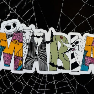 May include: A white sticker with the name "MARIA" written in colorful patchwork letters. The letters are decorated with black and white stripes, spiderwebs, and a cartoon character with a stitched mouth.