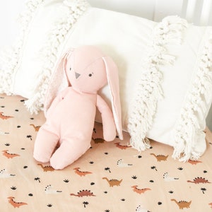 May include: A pink bunny soft toy with long ears sits on a tan cot sheet with a dinosaur print. The cot sheet has a pattern of brown dinosaurs and palm trees on a tan background.
