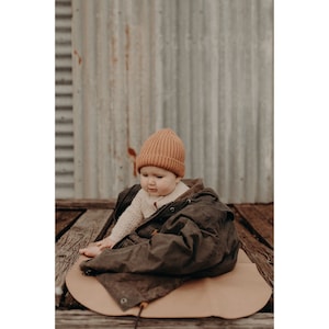 May include: A baby wearing a brown jacket and a rust-coloured beanie sits on a tan play mat. The baby is looking down at the mat.