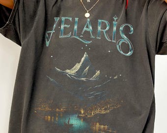May include: A charcoal gray short-sleeve t-shirt featuring the text VELARIS in a stylized font above a graphic illustration of a mountain landscape and a harbor at night with the text City of Starlight printed below the image.