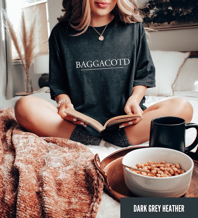 May include: A dark grey heather t-shirt with the text "BAGGACOTD" printed in white on the front. The shirt is being worn by a person sitting on a bed with a blanket, a book, a bowl of cereal, and a mug.
