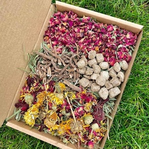 May include: A cardboard box filled with dried herbs and flowers for small animals. The box contains dried rose petals, dried calendula flowers, dried herbs, and small brown pellets.